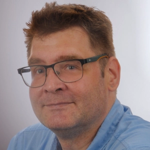 Martin von Pobrucki sitzt vor einem grauen Hintergrund und lächelt in die Kamera.
